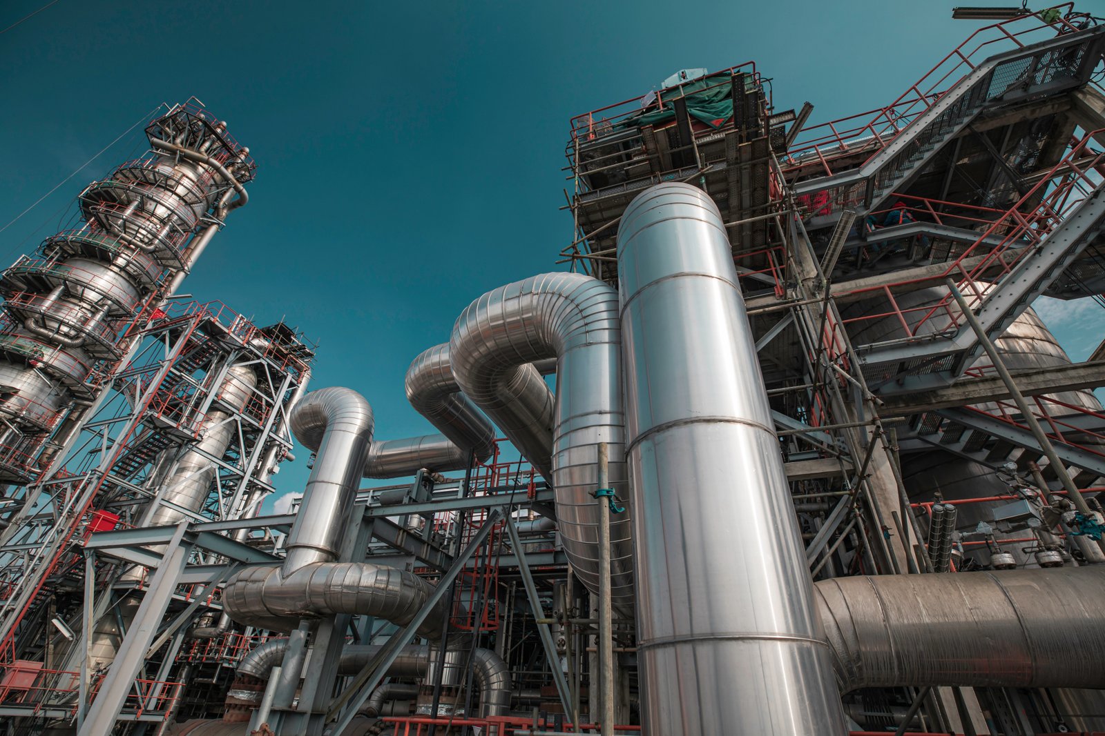 Industrial refinery in bright daylight with silver pipes, vessels, and red structural framework against a clear sky